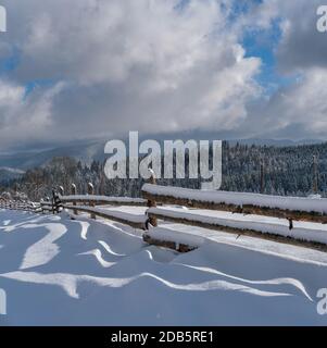 Pittoresche ombre ondate sulla neve da recinzione di legno. Borgo alpino di montagna invernale, sentiero innevato, bosco di abeti su colline molto nebbie e nuvolose. Foto Stock