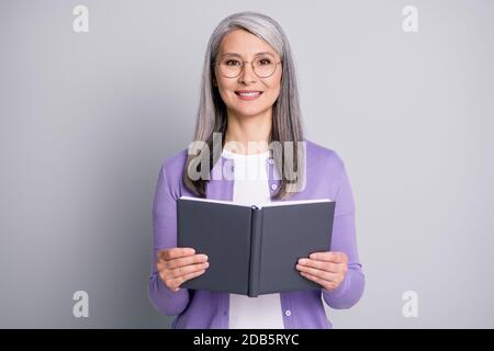Ritratto fotografico di una donna anziana che tiene un libro sorridente indossando occhiali cardigan viola isolato su sfondo grigio Foto Stock