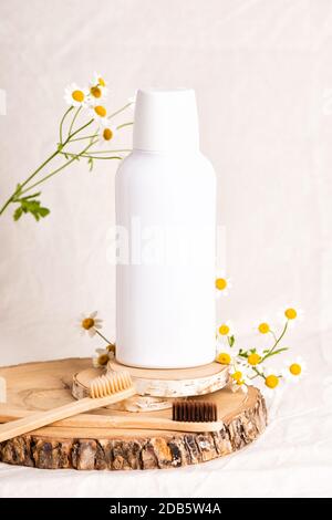 Bottiglia di prodotti cosmetici Mockup e spazzolini da denti in bambù con fiori di camomilla. Shampoo o collutorio su fetta di legno. Fondo in tessuto beige Foto Stock