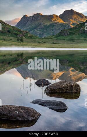 La luce del sole del mattino colpisce le vette montane con riflessi specchiati nel lago. Blea Tarn, Lake District, Regno Unito. Foto Stock