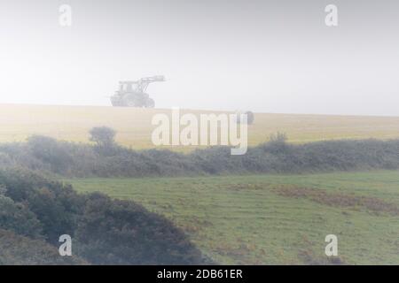 Un trattore che raccoglie balle rotonde di paglia in un campo in una giornata di nebbia in Cornovaglia. Foto Stock