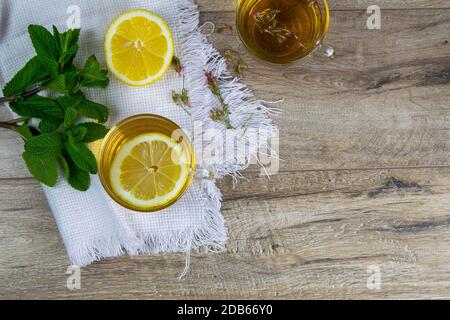 Tazza trasparente in vetro con tè alla menta su un supporto in legno, accanto a una ciotola di zucchero con cubetti di zucchero, foglie di limone e menta a fette. Foto Stock