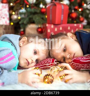 Una bambina nei vestiti domestici sta mentendo sotto l'albero di Natale vicino alle scatole con i regali sul cuscino e sui sogni. Luci di ghirlande, il befo notturno Foto Stock