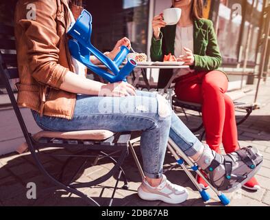 Due donne, una con una gamba rotta e stampelle, in un caffè con caffè e torta Foto Stock