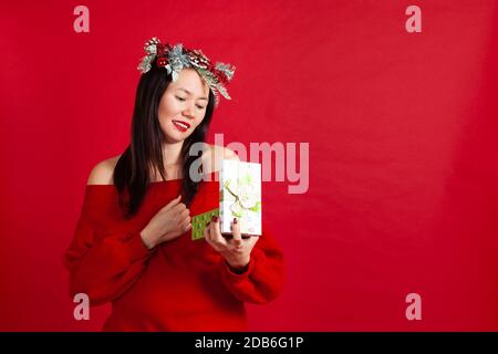 Mock up di bella giovane donna asiatica in corona di Natale con rossetto rosso apre un regalo di nuovo anno, su uno sfondo rosso Foto Stock