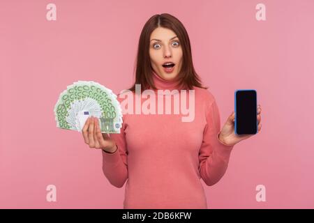 Donna bruna estremamente sorpresa in felpa rosa che tiene smartphone con schermo vuoto e fan di euro guardando fotocamera con espressione scioccata. Poll Foto Stock