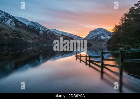 Recinzione allagata che conduce al lago con bei riflessi calmi di montagne innevate e vibrante alba arancione. Foto Stock