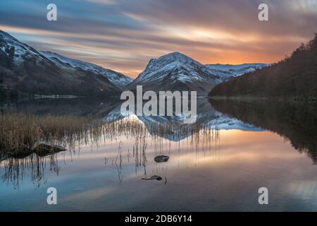 Vibrante alba arancione invernale in una tranquilla mattinata con riflessi di montagna innevati a Buttermere, nel Distretto dei Laghi Inglesi. Foto Stock