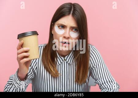 Donna stanca esausta con capelli marroni in camicia a righe bere caffè da tazza di carta seduta con macchie sotto gli occhi, alla ricerca di energia e motivati Foto Stock