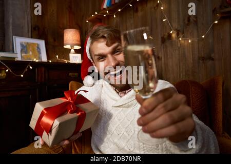 Ragazzo felice che indossa il cappello di santa che tiene il regalo e il bicchiere di champagne guardando la web cam. Foto Stock