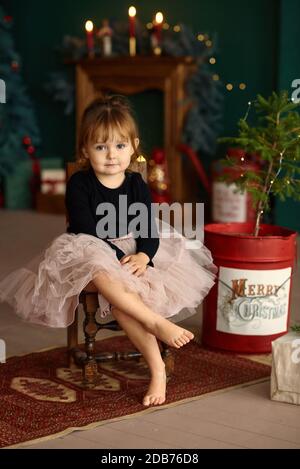 Ritratto di Natale di una bambina seduta su uno sgabello in una stanza con decorazioni natalizie. Ragazza carina vestita con gonna a tulle si posa in un elegante roo Foto Stock