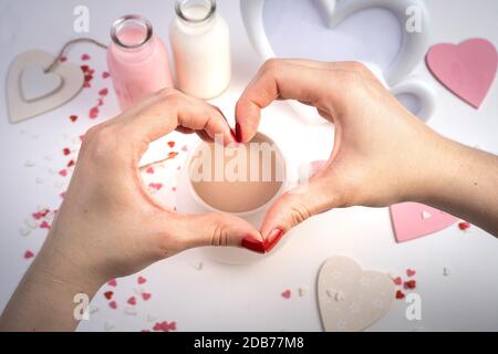 Mani di una donna a forma di cuore su uno sfondo romantico con una tazza di caffè con latte e cuori. Foto Stock