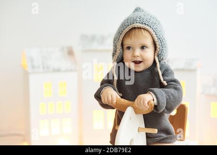 Un bambino piccolo in un cappello lavorato a maglia corre su un cavallo di legno in una stanza luminosa. Foto Stock