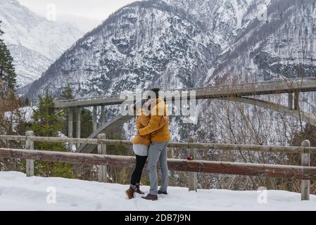 Una felice coppia baciante in cappotti coordinati su una neve giornata invernale con le montagne sullo sfondo Foto Stock