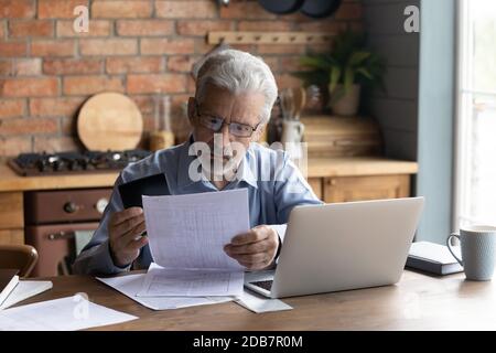 Uomo serio maturo che indossa occhiali calcolando le fatture domestiche, usando il laptop Foto Stock