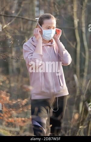 Ritratto della donna sportiva caucasica che indossa la maschera facciale di protezione medica mentre cammina nel parco, rilassandosi e ascoltando la musica. Corona virus, o Covid Foto Stock