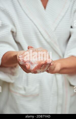 Donna in accappatoio bianco lavando le mani con schiuma di sapone, tagliato. Concetto di bellezza, cura della pelle e cosmetologia Foto Stock