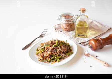 Porridge vegetariano sano di grano saraceno con fagioli verdi e carote, grano saraceno in un vaso di vetro, una bottiglia con olio su un tovagliolo beige, sale e pepe su Foto Stock