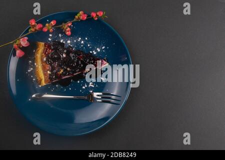 Fette di torta mousse di mirtillo con riflessi frutti di bosco decorati e foglie di menta su un piatto blu con fiori rossi sono su tavola nera, primo piano. Foto Stock