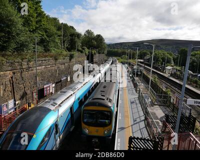 Due treni ferroviari blu giallo e argento che passano alla ferrovia stazione alto muro di pietra e alberi a sinistra stazione piattaforma a destra colline sullo sfondo Foto Stock