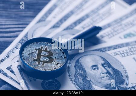 Bitcoin invece di lente di ingrandimento sullo sfondo di centinaia di dollari di bollette. Tonalità blu. Il concetto di sostituire il denaro della carta con quello virtuale Foto Stock