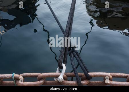 Dettaglio di due funi di ormeggio che attraversano prima di stringere una grande catena nel molo e riflessi di yacht di lusso in acqua. Foto Stock