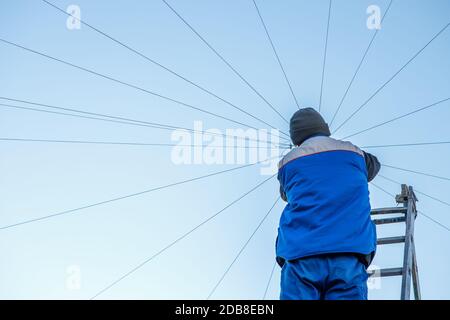Elettricista ripara il cablaggio elettrico sul tetto di un alto edificio che si erge sulle scale contro il cielo blu. Spazio di copia Foto Stock