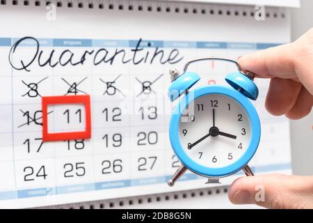 La mano tiene una piccola sveglia sullo sfondo del calendario del muro con l'iscrizione quarantena Foto Stock