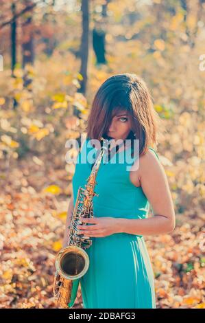 affascinante giovane ragazza bruna in un lungo abito blu gioca il sassofono alto in un parco autunnale giallo Foto Stock