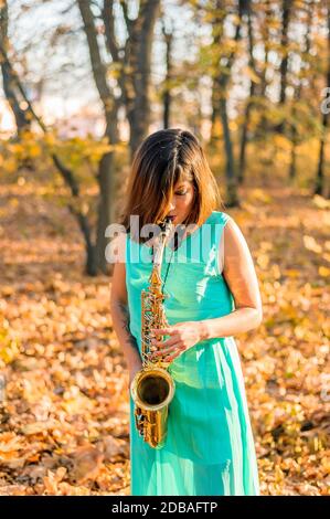 molto bella giovane ragazza bruna in un lussuoso abito blu lungo gioca solo sul suo sassofono nella foresta d'autunno Foto Stock