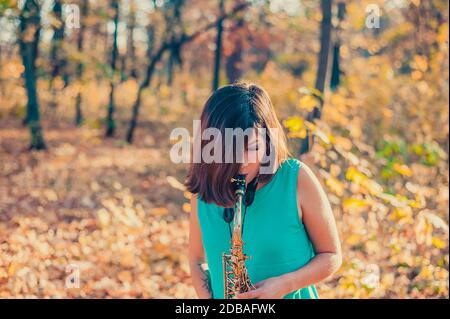 ragazza adolescente con capelli neri in un abito blu gioca con entusiasmo il sassofono nella foresta gialla di autunno Foto Stock