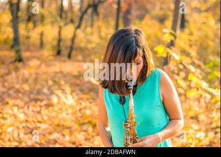 Giovane ragazza bruna con i capelli che coprono il suo viso gioca il sassofono, in piedi in abiti blu in un parco autunnale giallo Foto Stock