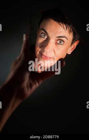 Ritratto facciale di una donna dai capelli neri di mezza età che guarda nella macchina fotografica in uno specchio a mano che brulica su uno sfondo scuro. Foto Stock