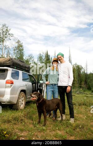 USA, Utah, Uinta National Park, Ritratto di coppia sorridente con il cane in piedi in prato, fuoristrada in background Foto Stock