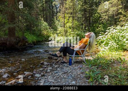 USA, Idaho, Sun Valley, Donna seduta sulla sedia lungo il fiume nella foresta Foto Stock