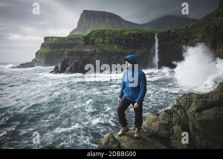 Danimarca, Isole Faroe, Gasadalur villaggio, Mulafossur cascata, uomo in piedi sulla scogliera con Mulafossur cascata in background Foto Stock