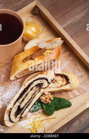 Lo strudel di papavero è decorato con zucchero in polvere, cannella, noci e menta e tè in una tazza di carta. Torta fatta in casa con ingredienti naturali. Legno rustico Foto Stock