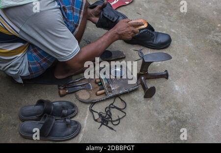 Indiano locale acciottolato riparazione scarpe accanto strada a mano utilizzando gli attrezzi in modo tradizionale. Naturale colpo raffigurante lavoro quotidiano di un regolare acciottolo. Foto Stock