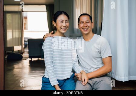 Ritratto di giovane coppia asiatica adulta abbracciando insieme con casa interiore in background. 30s felice marito e moglie matura sorridendo e guardando la macchina fotografica. Matrimonio e felice relazione vita concetto Foto Stock