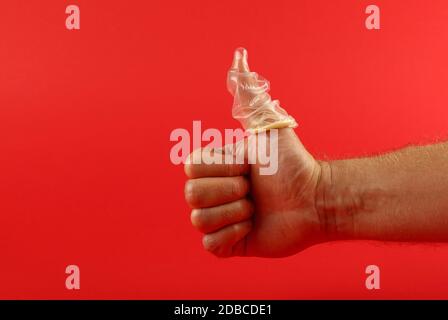 Close up man mano che mostra il pollice fino gesto con lattice preservativo sul dito su sfondo rosso con copia spazio, basso angolo vista laterale Foto Stock
