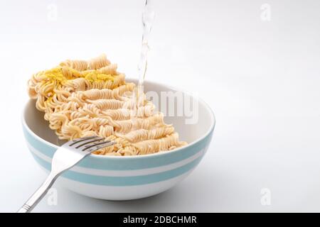 Alimenti a base di sodio elevato, spuntini veloci e pasti in pochi minuti con acqua calda che versa su tagliatelle istantanee in una ciotola in ceramica isolata su sfondo bianco Foto Stock