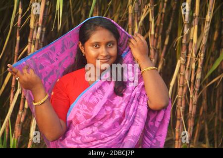 Primo piano di una ragazza indiana bengalese che indossa saree rosa e blusa rossa con orecchini color oro, bangle e capelli scuri che tengono sari sulla testa Foto Stock