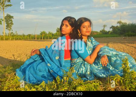 Primo piano di due ragazze adolescenti che indossano sari blu, orecchini, collana, con lunghi capelli scuri seduta indietro su un campo agricolo Foto Stock