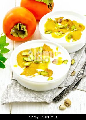 Dessert di yogurt, persimmon e miele con vaniglia, cardamomo e pistacchi in due ciotole su asciugamano, menta e cucchiaio su fondo di legno Foto Stock