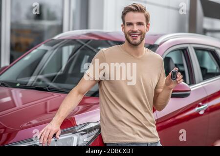 Sorridente giovane maschio appoggiato su auto rossa, tenendo le chiavi Foto Stock