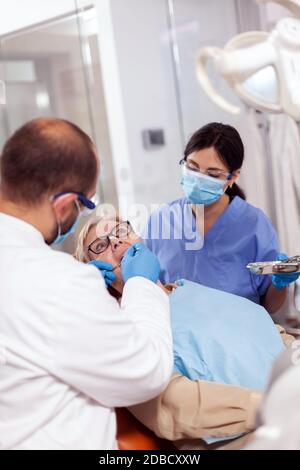 Dentista con assistente installare l'impianto nella clinica moderna della moutina del paziente. Donna anziana durante l'esame medico con stomatolog in studio dentistico con attrezzature arancioni. Foto Stock