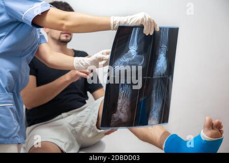 Medico femminile in un abito medico blu spiega ad un paziente di sesso maschile con una gamba rotta il risultato di una radiografia. Foto Stock