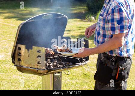 Un uomo alla griglia di carbone sta grigliando le bistecche, le salsicce alla griglia e le ali di pollo e usando le pinze del barbecue. Messa a fuoco selettiva sulla griglia contro una grana Foto Stock