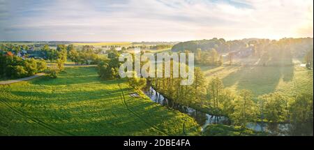 Primavera mattina panoramica rurale aerea. Alba su campi di colza verde e giallo fioriti. Nuvole di nebbia e piccolo fiume con alberi sulla riva del fiume. Bela Foto Stock