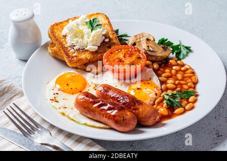 Colazione inglese con salsicce, fagioli, uova fritte, toast, funghi e pomodori in un piatto bianco. Concetto di colazione inglese. Foto Stock